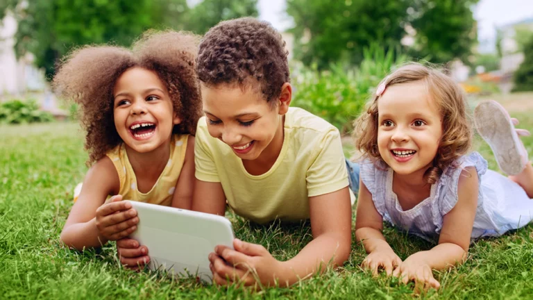 Three children happily using a tablet while lying on the grass, illustrating how screen time can be balanced with outdoor play.